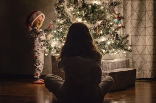 The baby is standing near the Christmas tree, the mother is sitting on the floor.