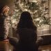The baby is standing near the Christmas tree, the mother is sitting on the floor.